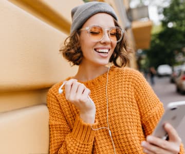 smiling girl in a sweater, glasses and a hat who holds a smartphone and headphones in her hands